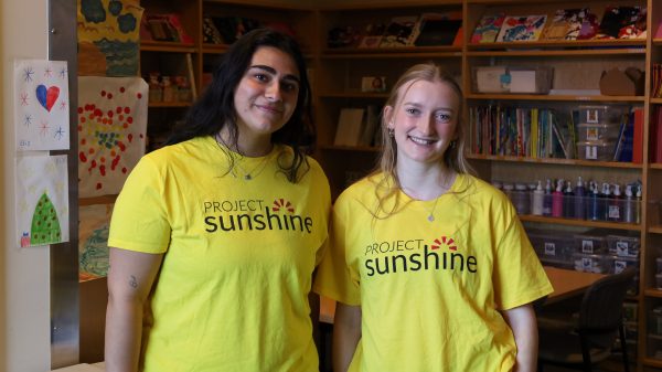 The image shows two individuals standing side by side in a room filled with shelves containing books, art supplies, and various artworks. Both individuals are wearing bright yellow T-shirts with the text "PROJECT SUNSHINE" printed on them. The background shows a well-organized space with tables and chairs, suggesting an environment conducive to creative or educational activities.