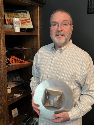Person wearing a light plaid shirt holding a large metallic geometric object, standing in front of a wooden shelf displaying Syracuse memorabilia and various items