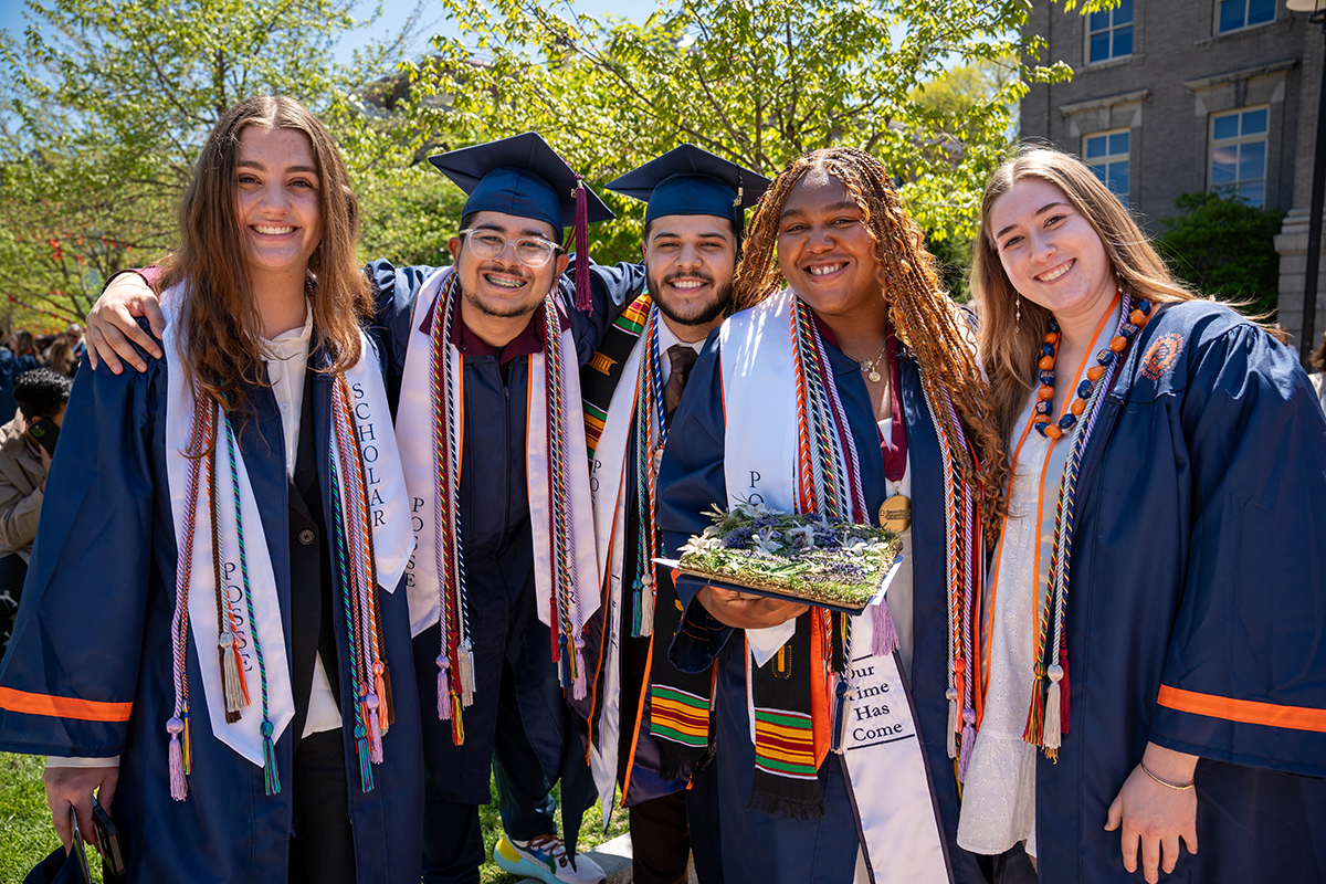 A group of five people dressed in graduation gowns and caps, standing outdoors. They are wearing various cords and stoles, indicating academic achievements and affiliations. One person is holding a decorated graduation cap. The background includes trees and a building.