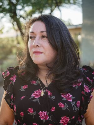 The image shows a person with shoulder-length dark hair wearing a black dress adorned with pink and red floral patterns. They have a gold necklace featuring a pendant in the shape of an animal. The background is an outdoor setting with trees and sunlight filtering through, creating a serene atmosphere.