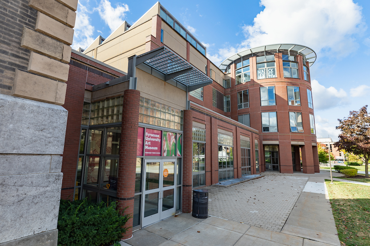 The image shows the exterior of a modern building with a mix of brick and glass architecture. The building has large windows, a curved section on the right side, and an entrance with glass doors on the left. Above the entrance, there is a sign that reads "Syracuse University Art Museum." The sky is partly cloudy, and there are some trees and shrubs around the building.