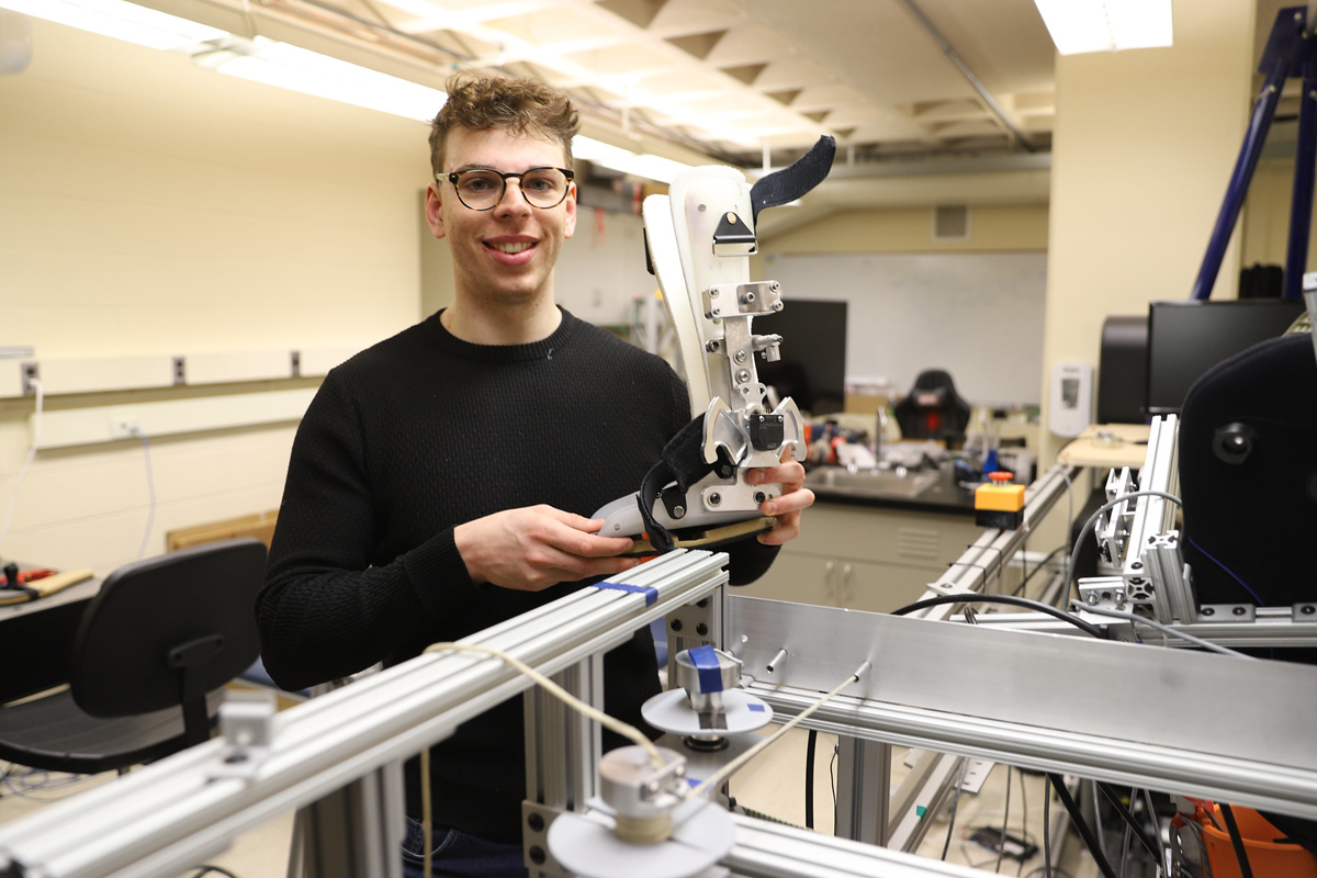 The image shows a person holding a mechanical device in what appears to be a laboratory or workshop setting. The device seems to be part of an engineering or robotics project, and the room contains various pieces of equipment and tools.