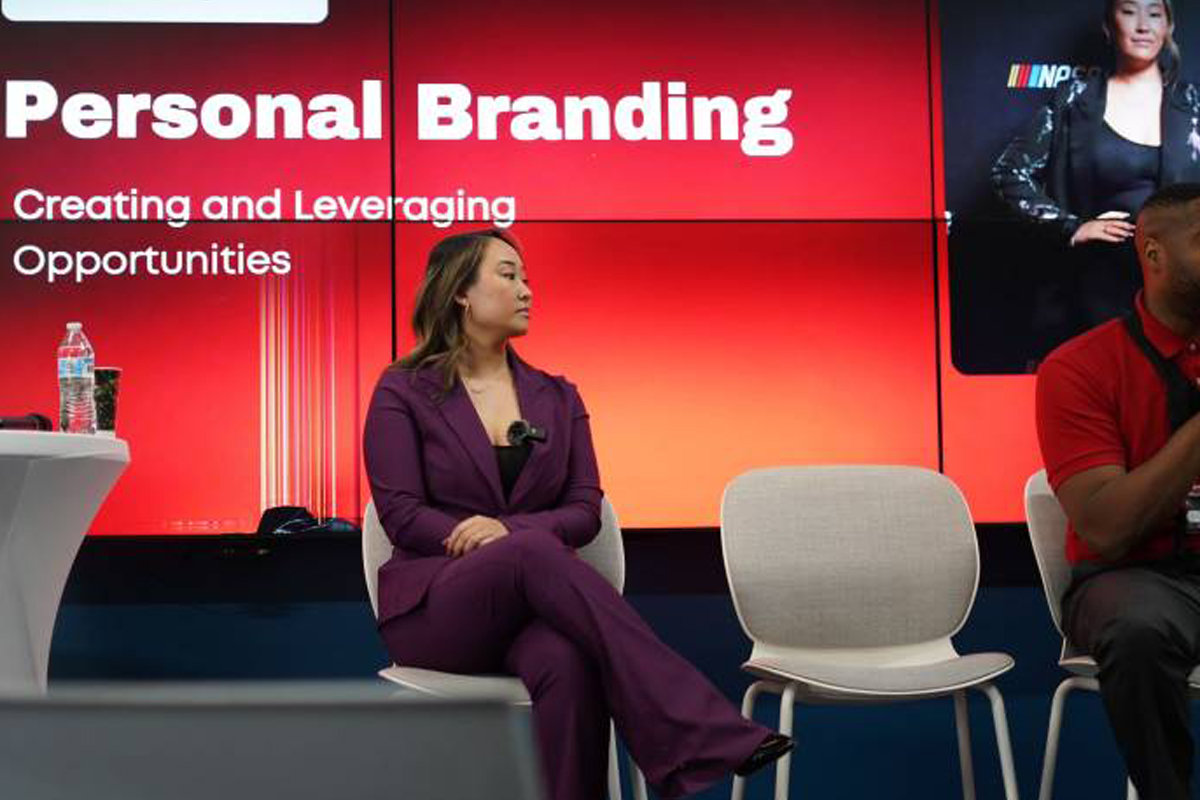 The image shows a panel discussion or presentation setting with three individuals seated on chairs. The background features a large screen displaying the text "2025 Personal Branding: Creating and Leveraging Opportunities." One individual is wearing a purple suit, sitting in the center, while another individual in a red shirt is seated to the right. A water bottle and some papers are placed on a small table to the left.