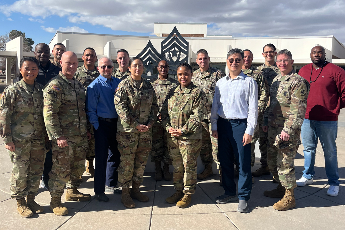 The image shows a group of people, some in military uniforms and others in civilian clothing, standing together outdoors. In the background, there's a building with a distinctive emblem on it. The sky is partly cloudy.