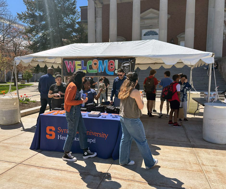 A group of people are gathered outside under a white canopy tent with a colorful 'WELCOME!' banner hanging at the back. The tent is set up in front of Hendrick's Chapel's large columns and steps. There is a table covered with a blue tablecloth that reads 'Syracuse University Hendricks Chapel' in orange and white text. Various items are displayed on the table, including pamphlets and small objects. Some people are standing near the table, while others are walking by or talking to each other. Trees and other buildings can be seen in the background.