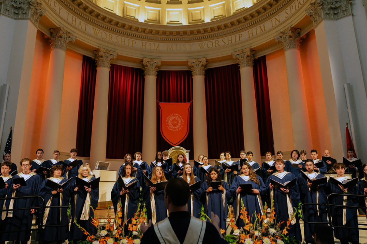 A choir performance in a grand hall with tall columns and red curtains. The choir members, dressed in blue robes, are standing on risers and singing. In the foreground, a conductor faces the choir. The backdrop features an orange banner with an emblem, and above it, an inscription reads "WORSHIP HIM THAT MADE HEAVEN AND EARTH AND THE SEA AND THE FOUNTAINS OF WATERS." Floral arrangements are placed at the base of the risers.