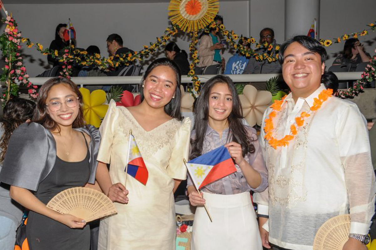 The image shows four people standing together, each holding a small Philippine flag. They are dressed in traditional Filipino attire. The background is decorated with colorful flowers and festive decorations, and there are more people visible behind them on an elevated platform.