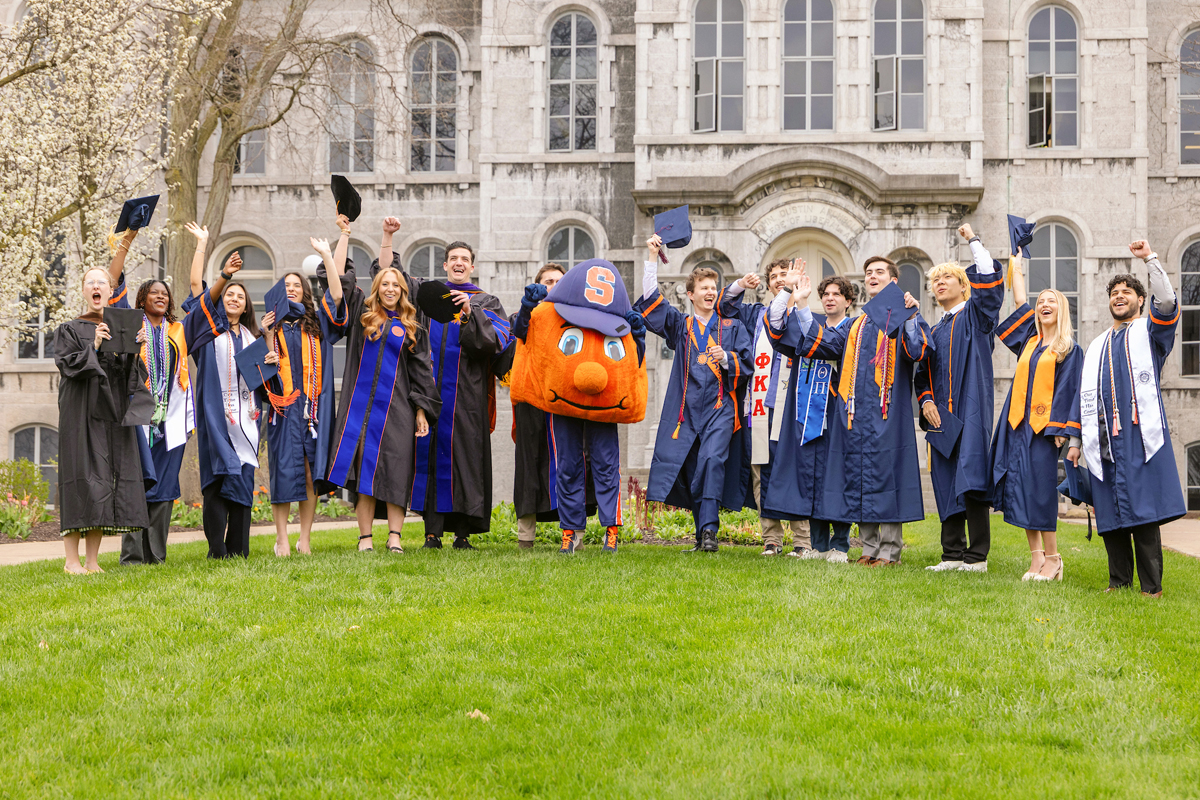 A group of people dressed in graduation gowns and caps standing on a grassy area in front of a large stone building. They are celebrating, with some holding their caps in the air and others raising their arms. In the center of the group is a person wearing an orange mascot costume with a blue cap that has an "S" on it. The building behind them has arched windows.