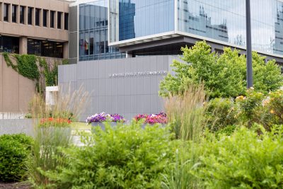 S.I. Newhouse School of Public Communications building with glass windows, surrounded by greenery and flower beds; ivy-covered building in the background.
