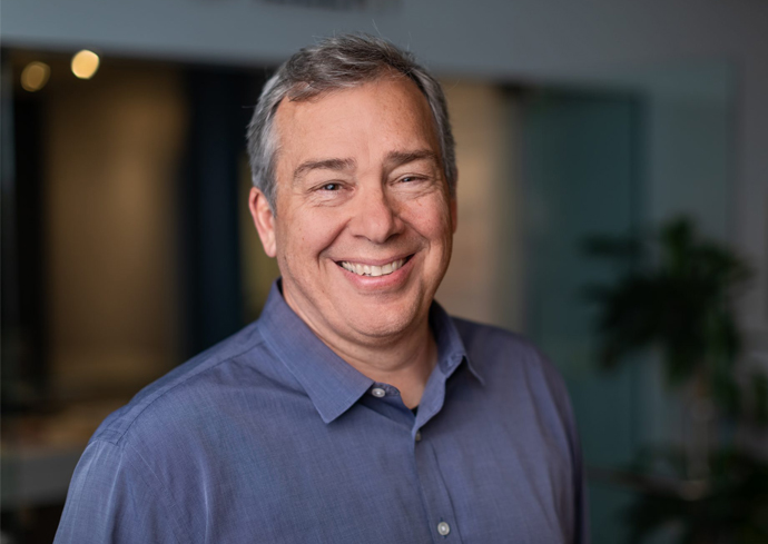 The image shows a person with short, gray hair wearing a blue button-up shirt. The background appears to be an indoor setting with blurred elements, including some lights and possibly plants.