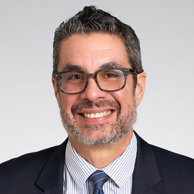 The image shows a person wearing a dark suit, white and blue striped dress shirt, and a blue tie with diagonal stripes. The person has short, dark hair and is wearing glasses. The background is plain and light-colored.