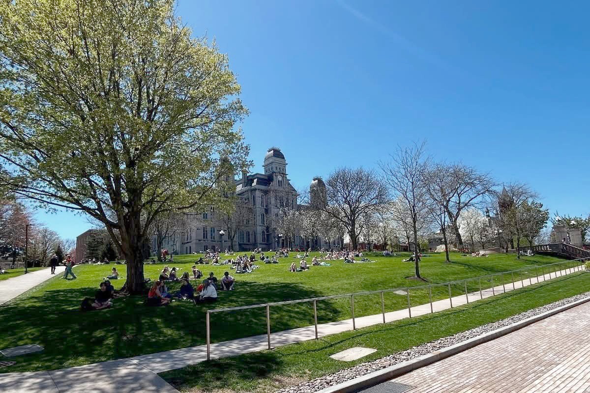 The image shows a large, grassy area on a university campus with several groups of people sitting and lying on the grass. In the background, there is an ornate, historic building with a dome and multiple spires. The sky is clear and blue, indicating a sunny day. There are trees around the grassy area, some with leaves and some without. A paved walkway runs through the grass, and there is a metal railing along one side of the path.
