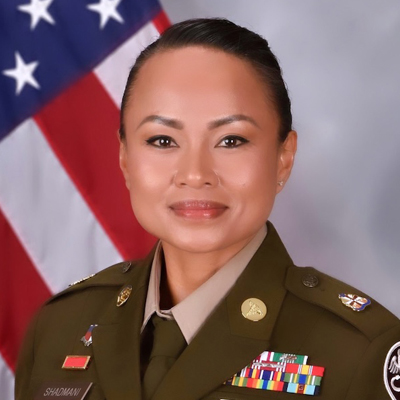 A military officer smiles while posing for a headshot in uniform. An American flag is in the background.