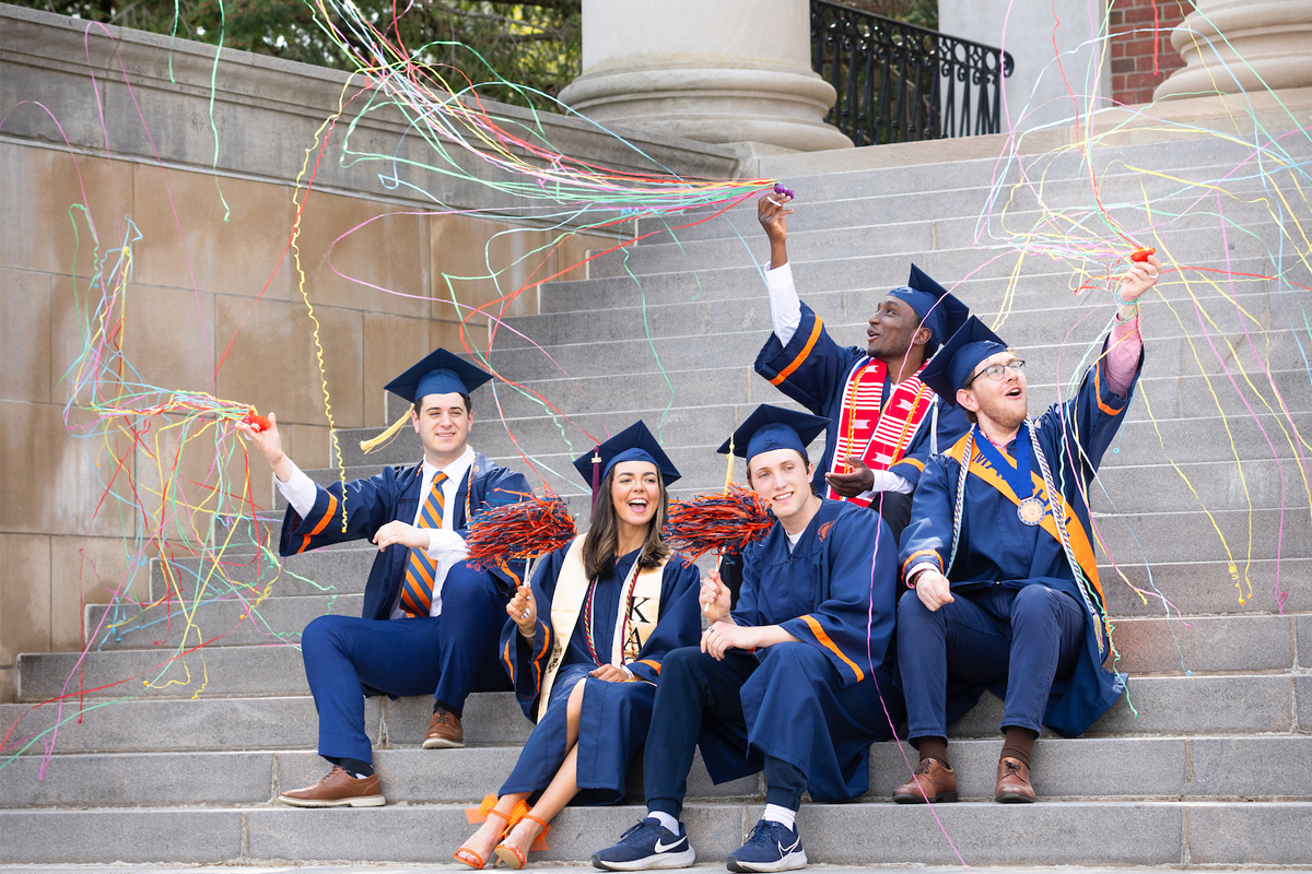 A group of five people dressed in graduation gowns and caps, sitting on the steps of a building. They are holding and waving colorful streamers, which are flying through the air around them. The background includes stone steps and columns.