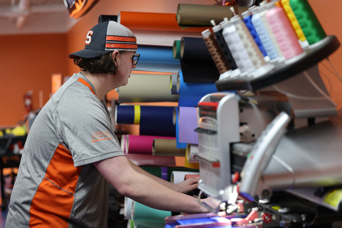 The image shows a person wearing a gray and orange shirt and a cap, operating a machine in what appears to be a print shop or workshop. The background features various rolls of colored vinyl or paper stacked on shelves. The machine the person is using has multiple spools of thread, indicating it might be an embroidery or printing machine.