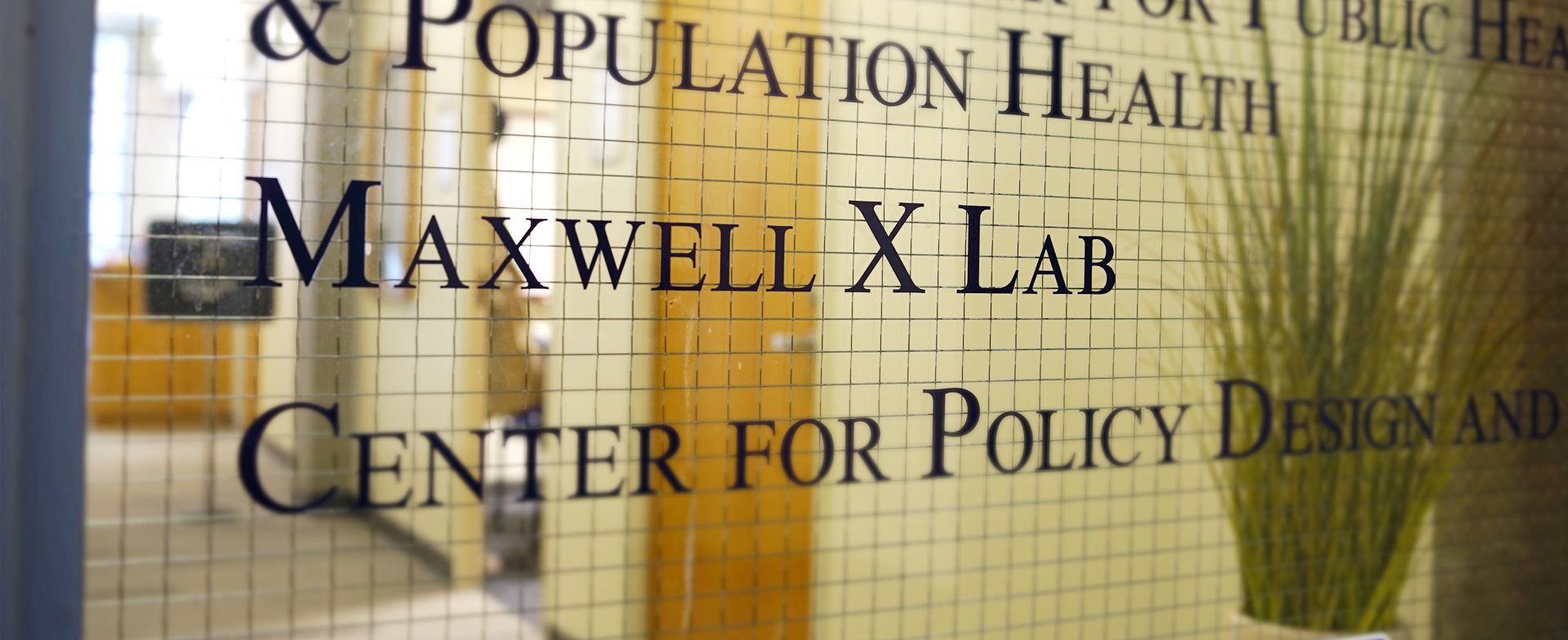 The image shows a glass door with a wire mesh pattern, leading to an office or lab. The text on the door reads: "MAXWELL X LAB CENTER FOR POLICY DESIGN AND POPULATION HEALTH." In the background, there is a hallway with wooden doors and a plant visible through the glass.