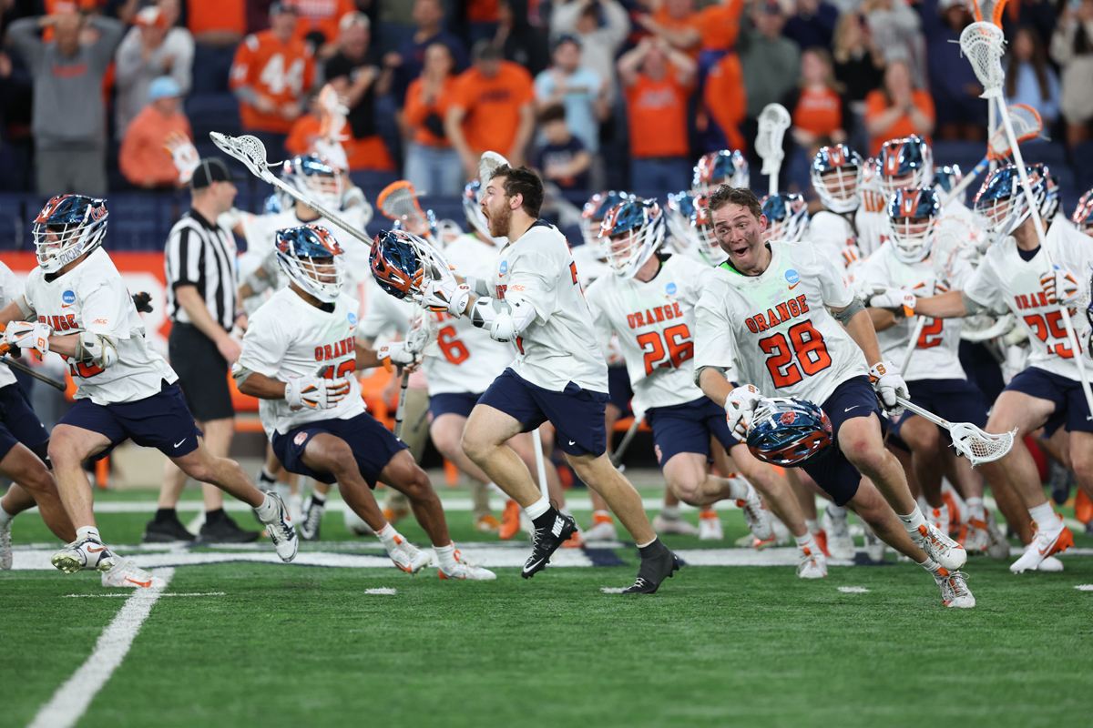 A group of lacrosse players in white jerseys with "ORANGE" written on them are running on a field. The players are wearing helmets and holding lacrosse sticks. There is a referee in the background, and spectators can be seen in the stands. The image captures the moment of action as the players move forward energetically.