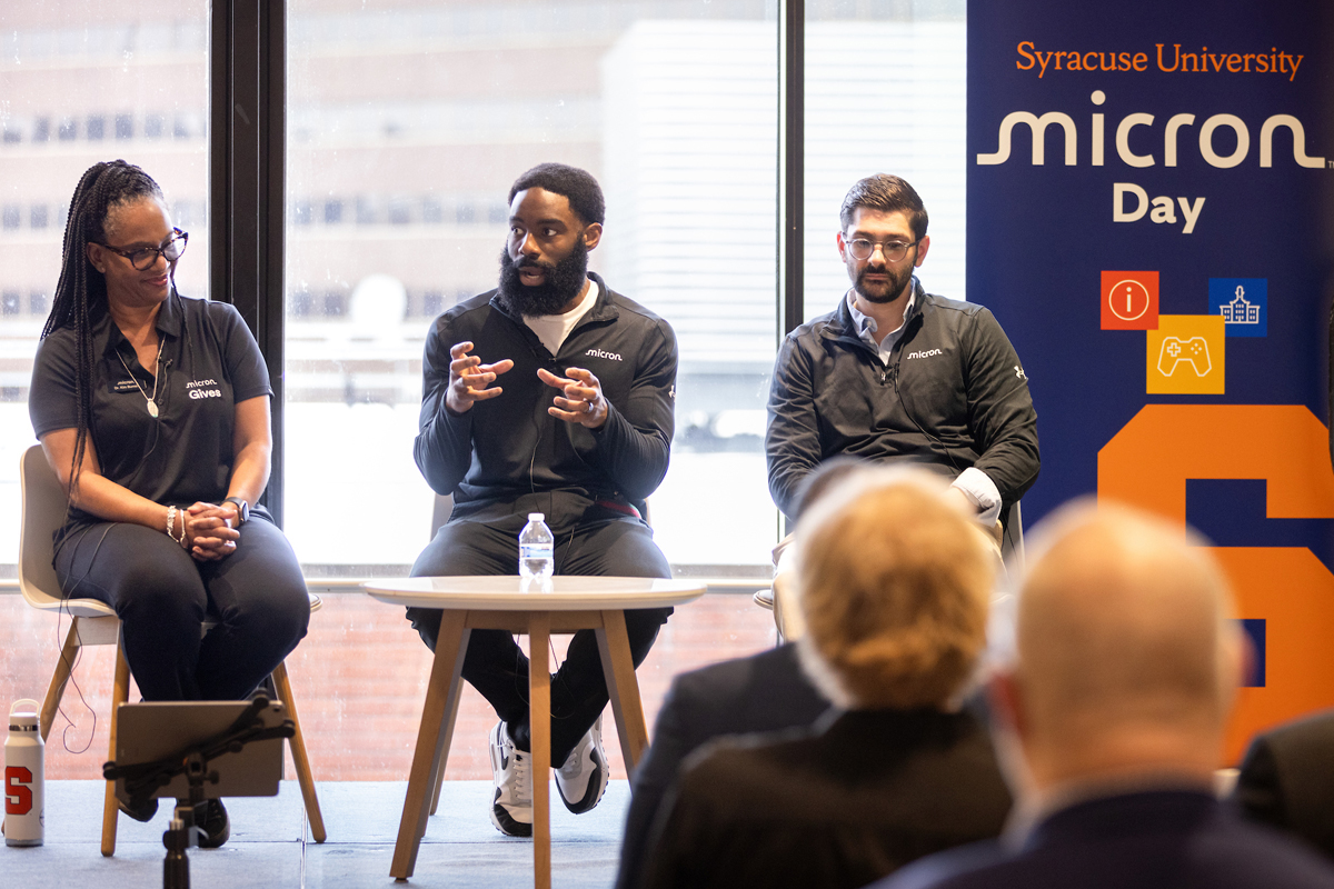 The image shows three individuals seated on a stage in front of an audience, participating in a panel discussion at an event called "Micron Day" hosted by Syracuse University. They are dressed in black attire with the Micron logo. 