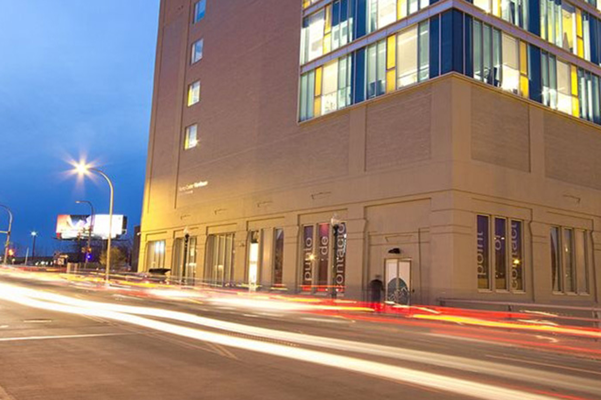 The image shows a modern multi-story building with large windows at dusk. It is located on a street corner, and the long exposure photograph captures light trails from passing vehicles, creating streaks of white and red lights along the road. The sky is a deep blue, indicating that it is early evening.