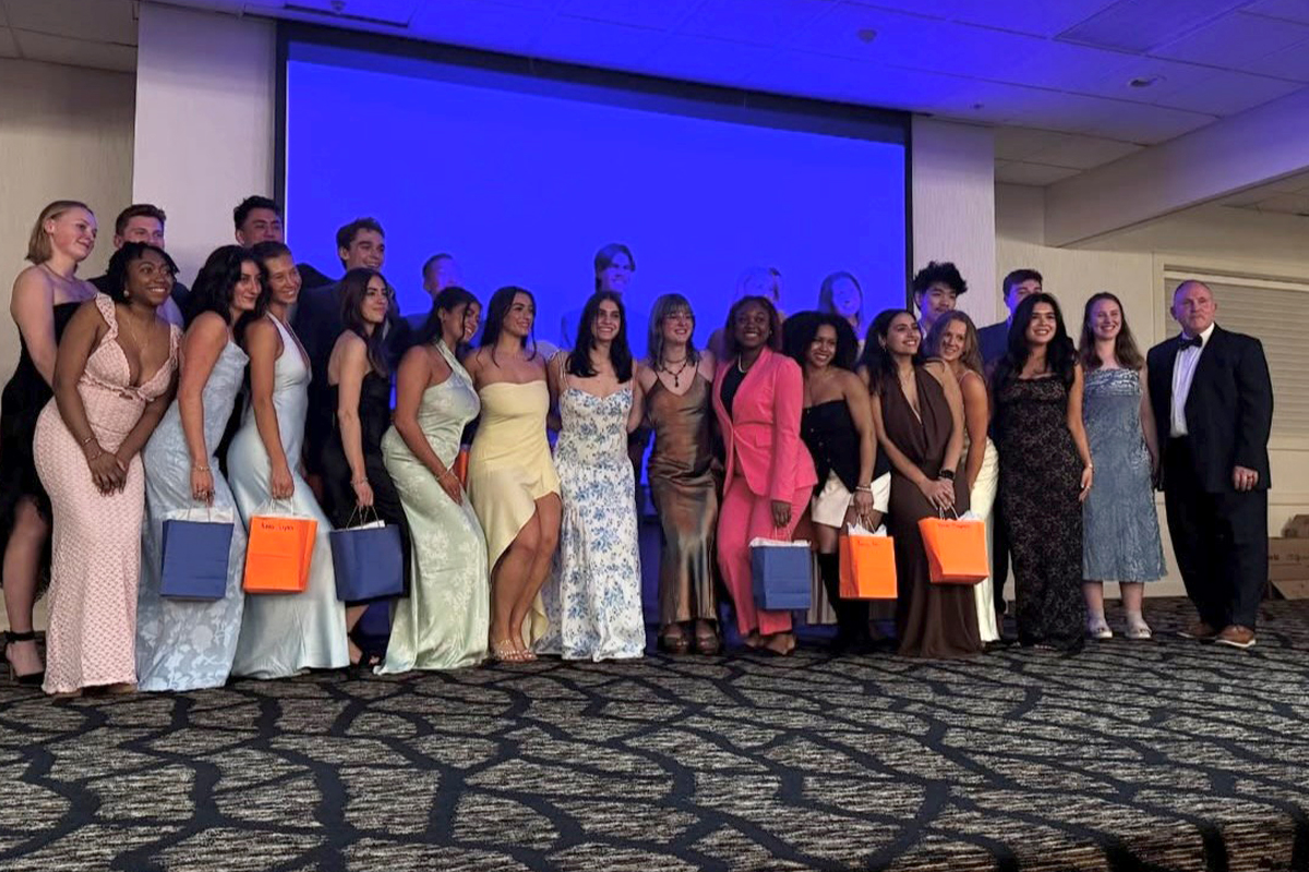 A group of people dressed in formal attire are standing together in a room with patterned carpet and a blue-lit background. Some individuals are holding gift bags, and everyone is posing for a group photo.