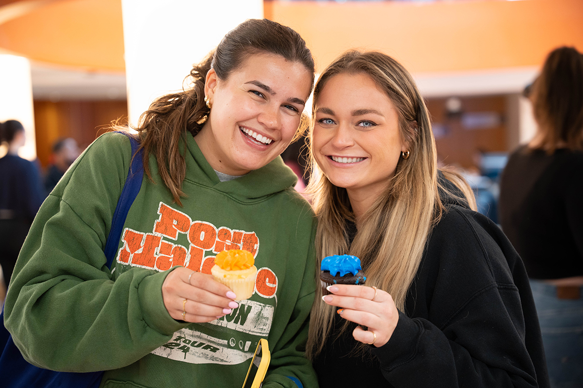 Two people standing close together, each holding a dessert. The person on the left is wearing a green hoodie and holds an orange-colored cupcake. The person on the right is wearing a black hoodie and is holding a blue-frosted cupcake.