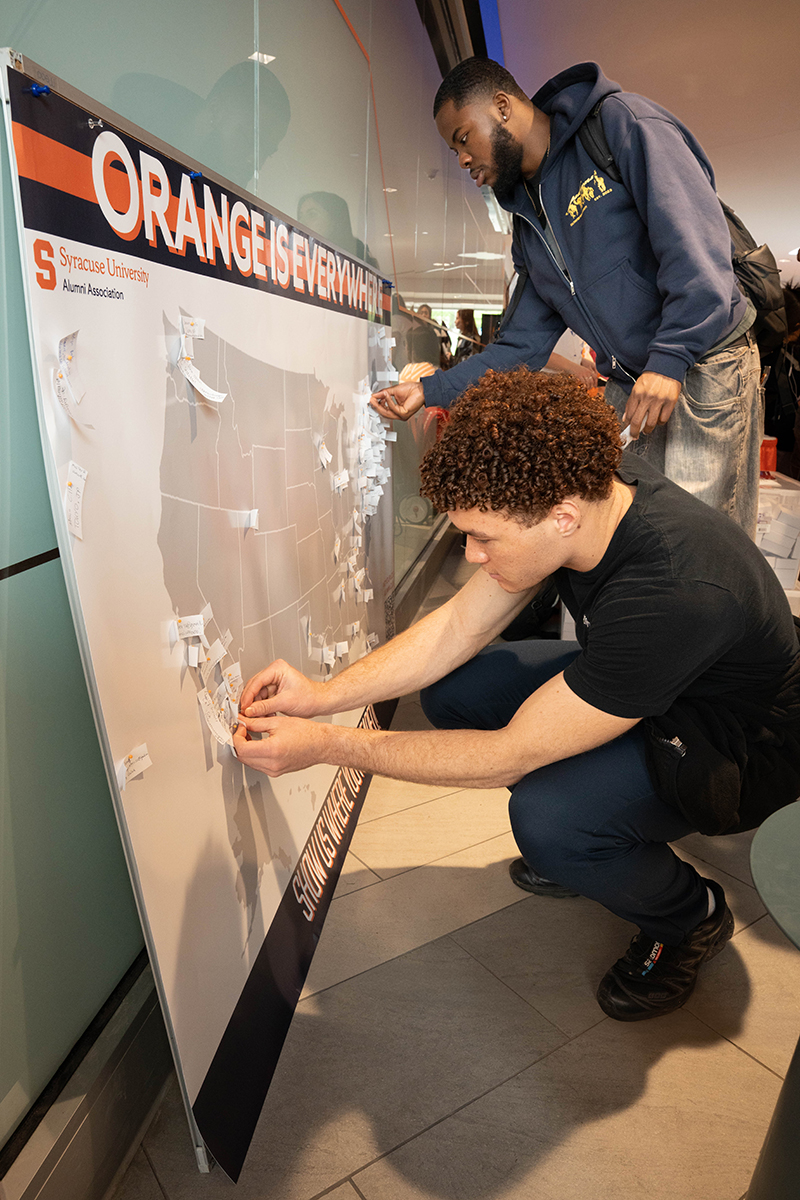 Two individuals place tags on a large map of the United States. The map is part of a display with the heading 'ORANGE IS EVERYWHERE' created by the Syracuse University Alumni Association.