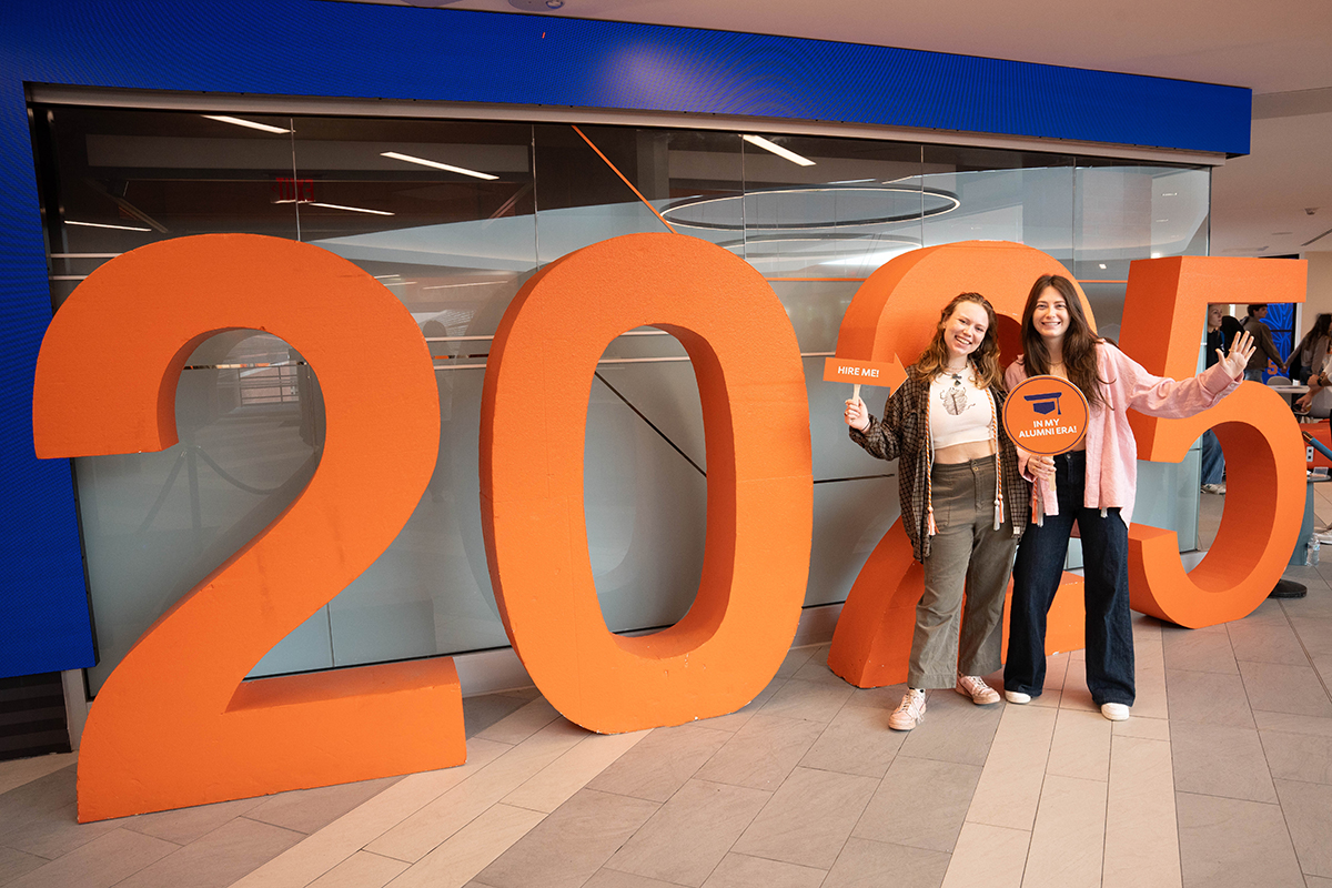 Two people stand in front of large orange numbers '2025'. The person on the left holds a sign that says "HIRE ME" and the person on the right holds a sign that says "IN MY ALUMNI ERA".