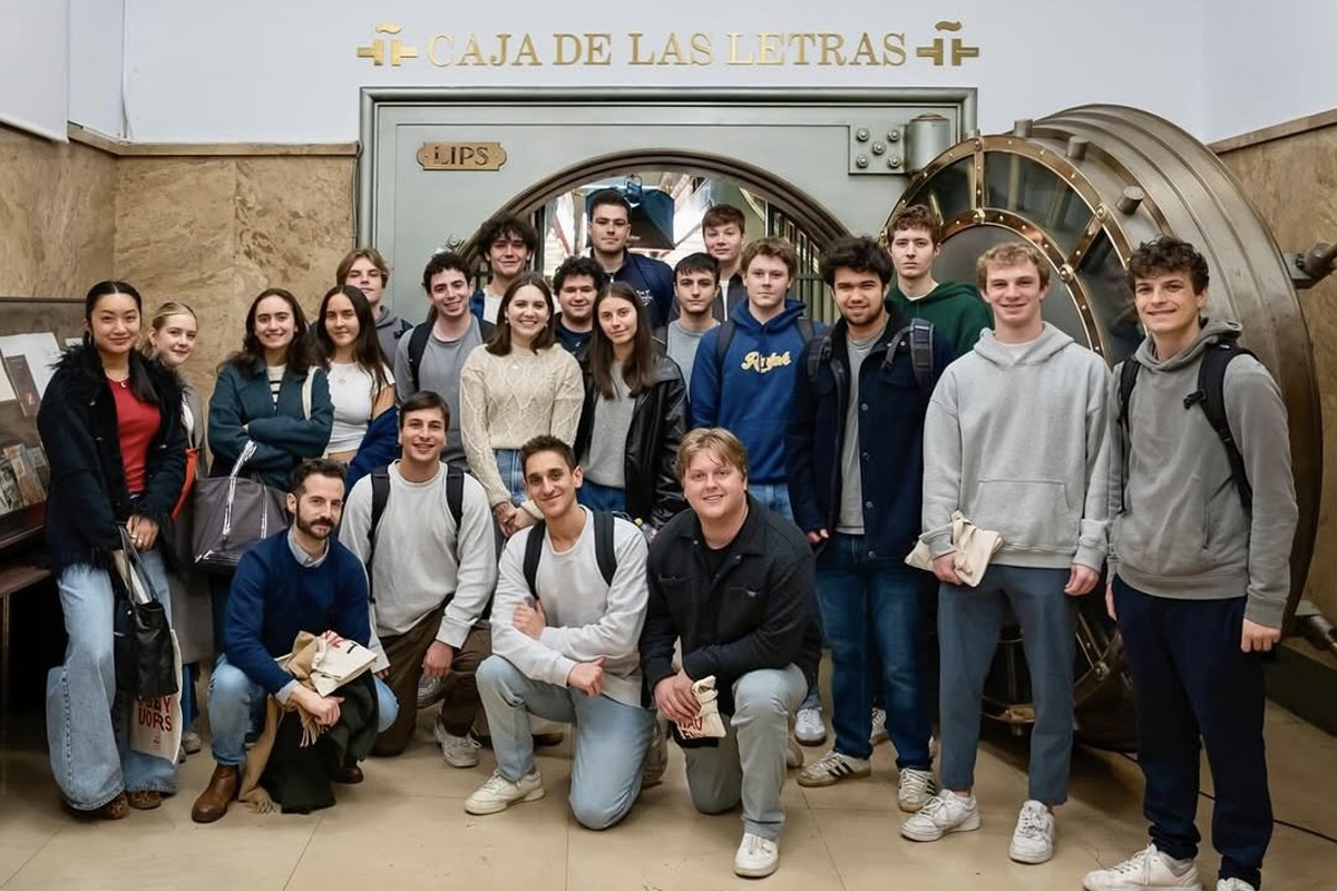 The image shows a group of people standing in front of a large vault door with the words "CAJA DE LAS LETRAS" written above it. The vault door has a circular design and appears to be made of metal. Some individuals are holding bags, and there is a display case on the left side of the image. The background includes marble walls and a tiled floor.