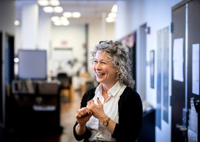 The image shows a person with curly gray hair standing in an office hallway. They are wearing a white shirt and a black cardigan, and appear to be gesturing with their hands. The background features an office environment with desks, chairs, and fluorescent lighting.