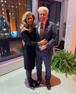 Smiling couple standing together indoors with city lights visible through large windows and a green fern nearby.