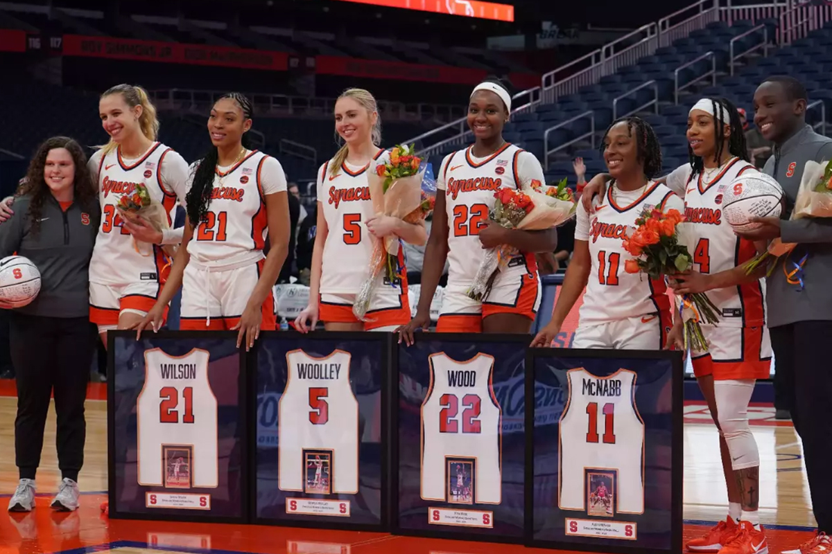 The image shows a group of basketball players standing in a row on a basketball court. Each player is holding flowers and standing behind framed jerseys with their names and numbers. The jerseys displayed are:Wilson - Number 21 Woodley - Number 5 Wood - Number 22 McNabb - Number 11