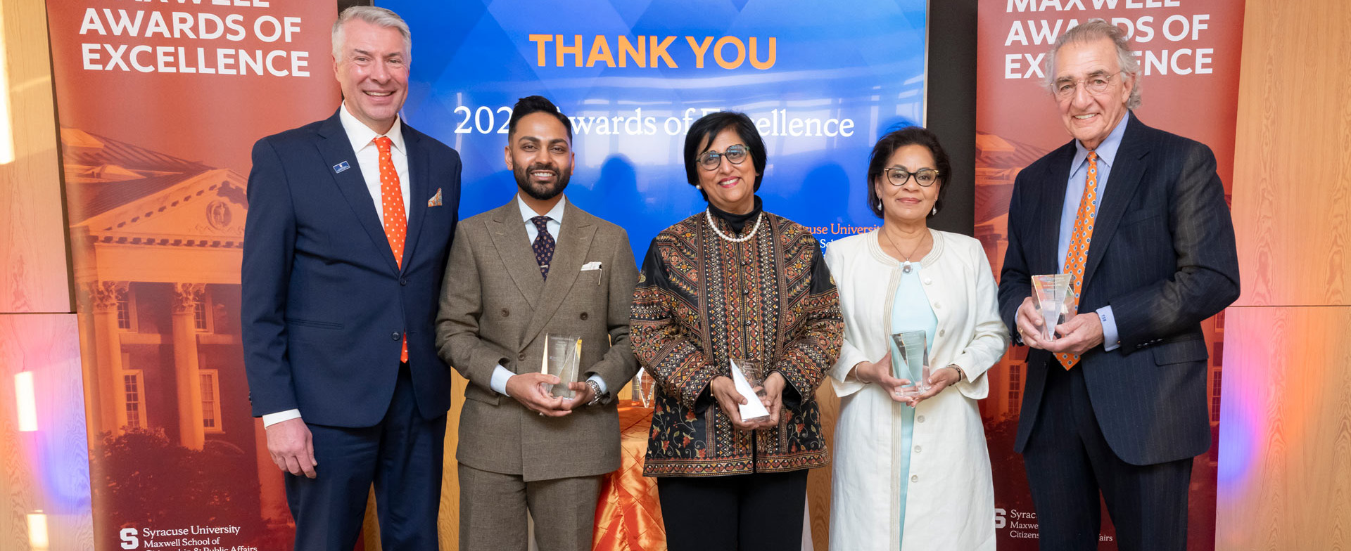 The image shows five individuals standing in front of a backdrop that reads "THANK YOU 2023 Awards of Excellence" and "MAXWELL AWARDS OF EXCELLENCE." Each person is holding an award. The backdrop also features the Syracuse University Maxwell School logo and an image of a building.