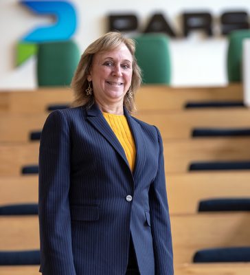 The image shows a person standing indoors. They are wearing a navy blue pinstripe blazer over a bright yellow top. The background features wooden steps and green cushioned seats, with part of a colorful abstract logo visible on the wall behind.