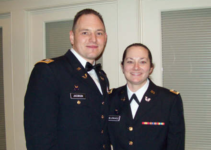 The image shows two individuals standing side by side, dressed in formal military uniforms. The individual on the left is wearing a black bow tie and has a name tag that reads "ANDERSON." The individual on the right has a name tag that reads "BLANAS" and is adorned with several colorful ribbons on the chest. They are standing in front of a set of closed doors with glass panels.