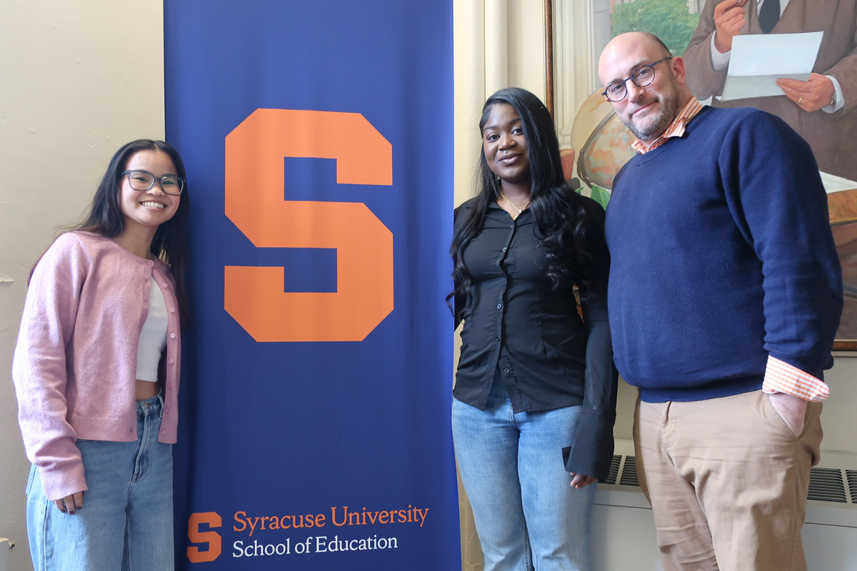 The image shows three people standing next to a blue banner with an orange "S" and the text "Syracuse University School of Education."
