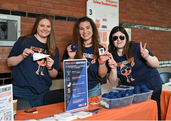 three people standing behind a table promoting Syracuse University Giving Day