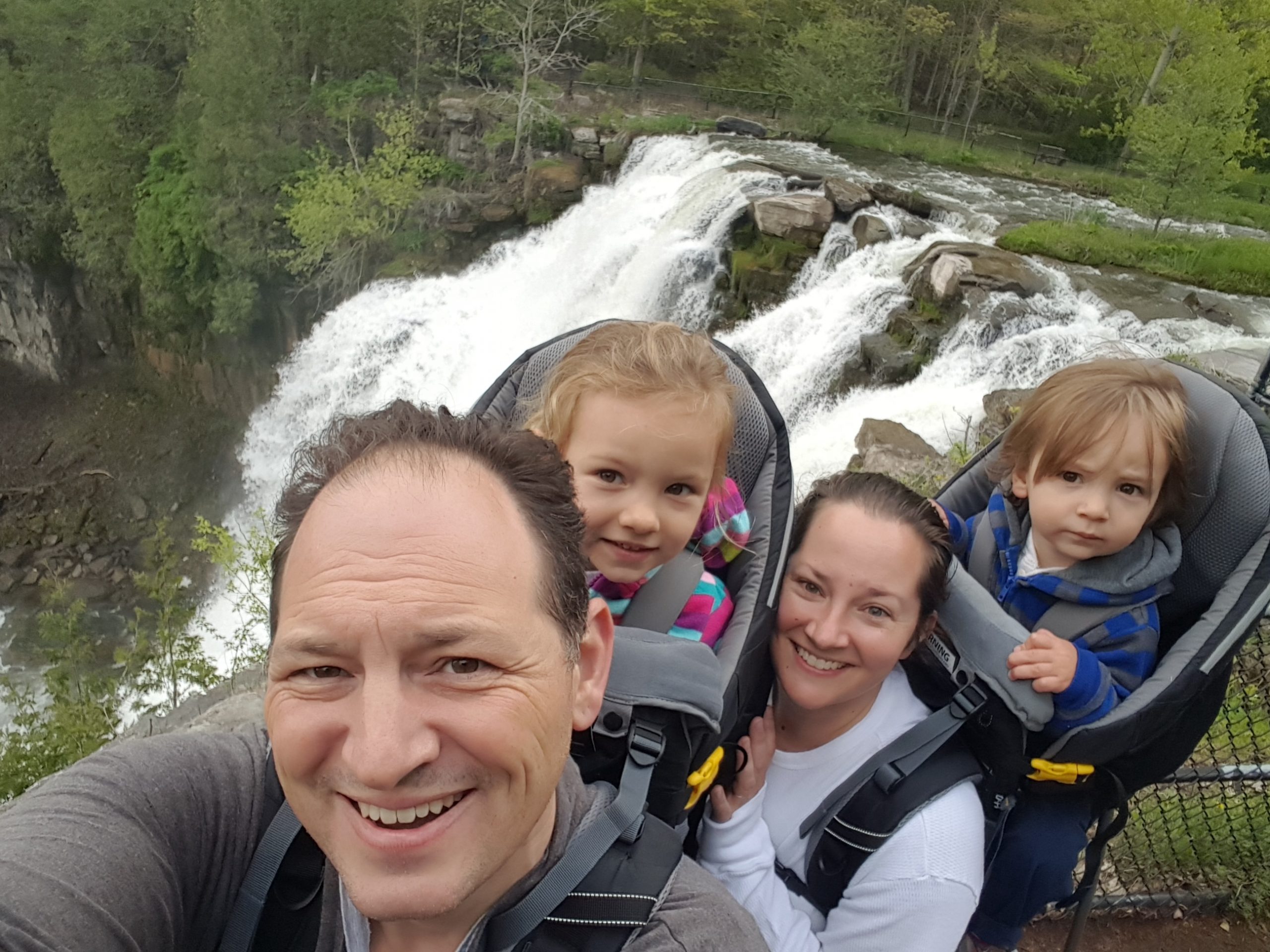 The image shows a group of people, including two adults and two children, standing in front of a waterfall. The adults are carrying the children in backpack carriers. The background features a lush green forest and the cascading waterfall.