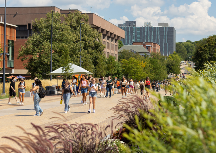 The image shows a busy college campus walkway with numerous students walking in various directions. The scene is vibrant with greenery, including trees and plants along the path. Several buildings are visible in the background, including a modern multi-story building and older structures. The sky is partly cloudy, adding to the bright and lively atmosphere of the campus.