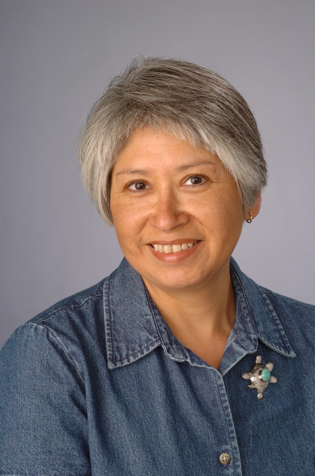 The image shows a person with short, gray hair wearing a denim shirt. The person's face is blurred out. There is a brooch pinned to the shirt on the left side of the chest. The background is plain and light-colored.