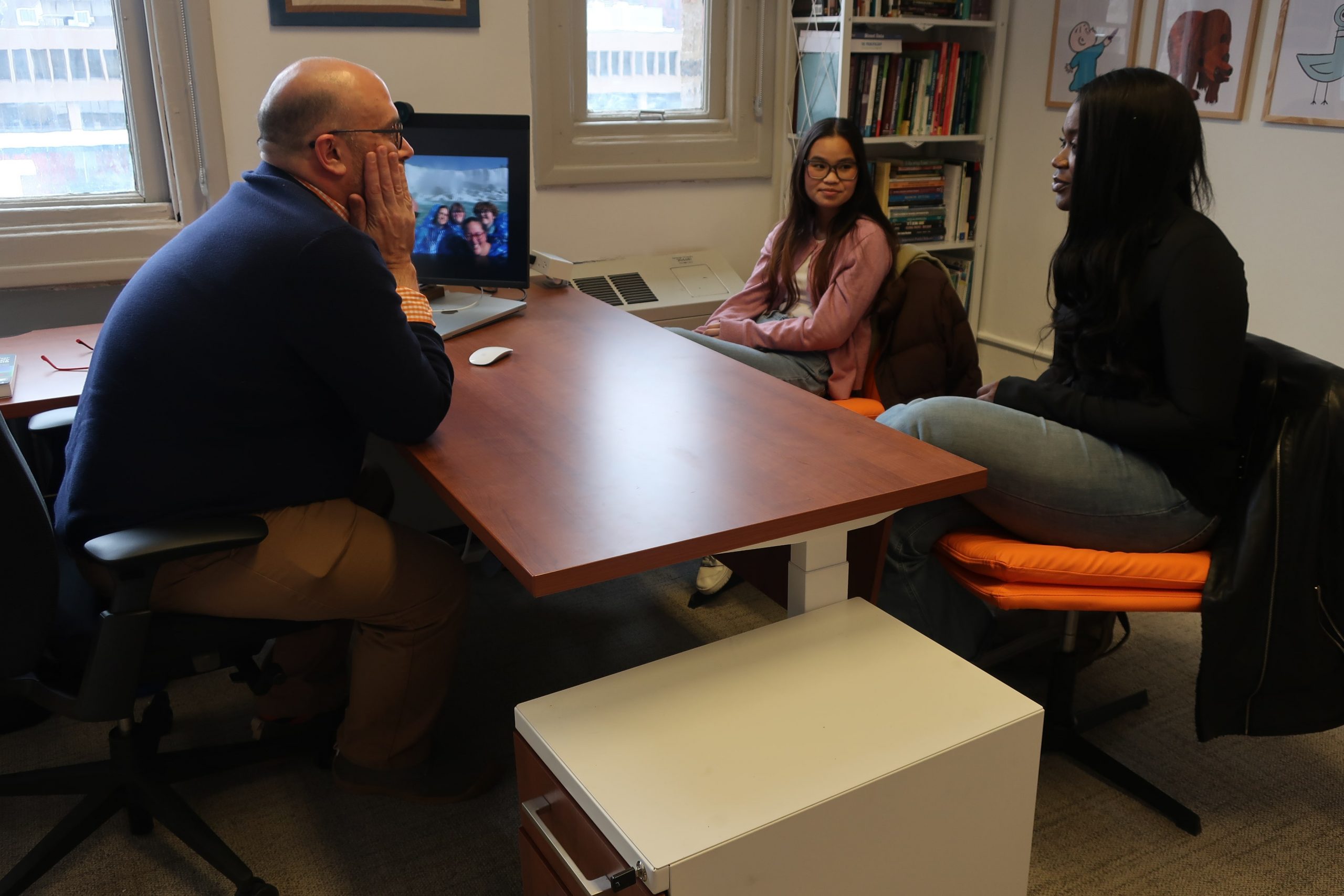 The image shows three people sitting in an office setting. One person is seated on the left side of a wooden desk facing two other individuals, who are seated on the right side.