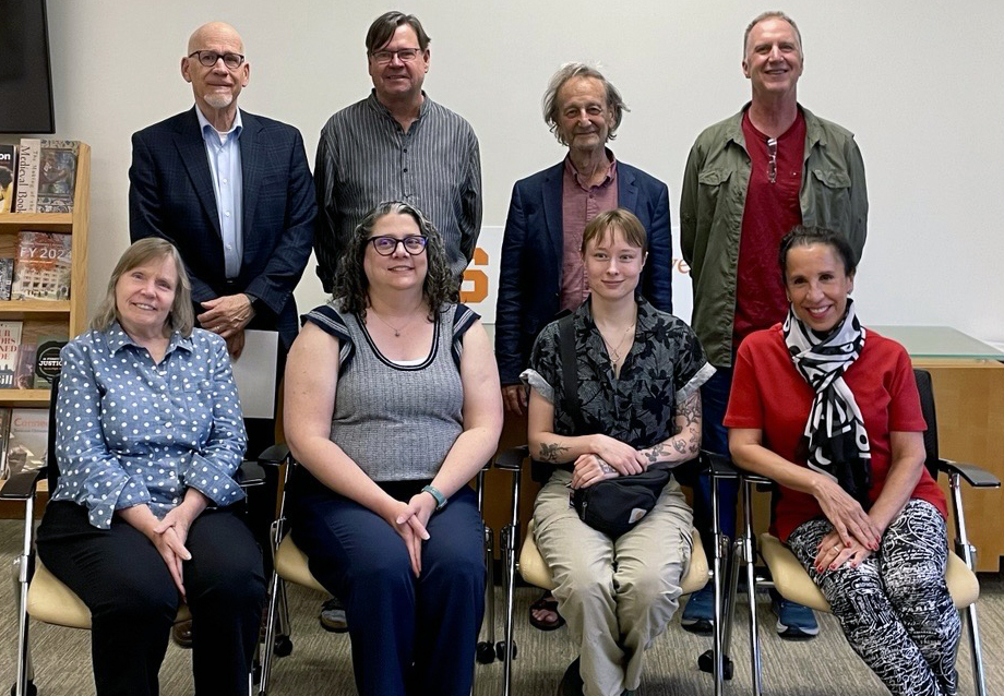 A group photo showing eight adults, four seated in the front row and four standing behind them. The background includes a bookshelf with various items and a white wall with partial text visible.