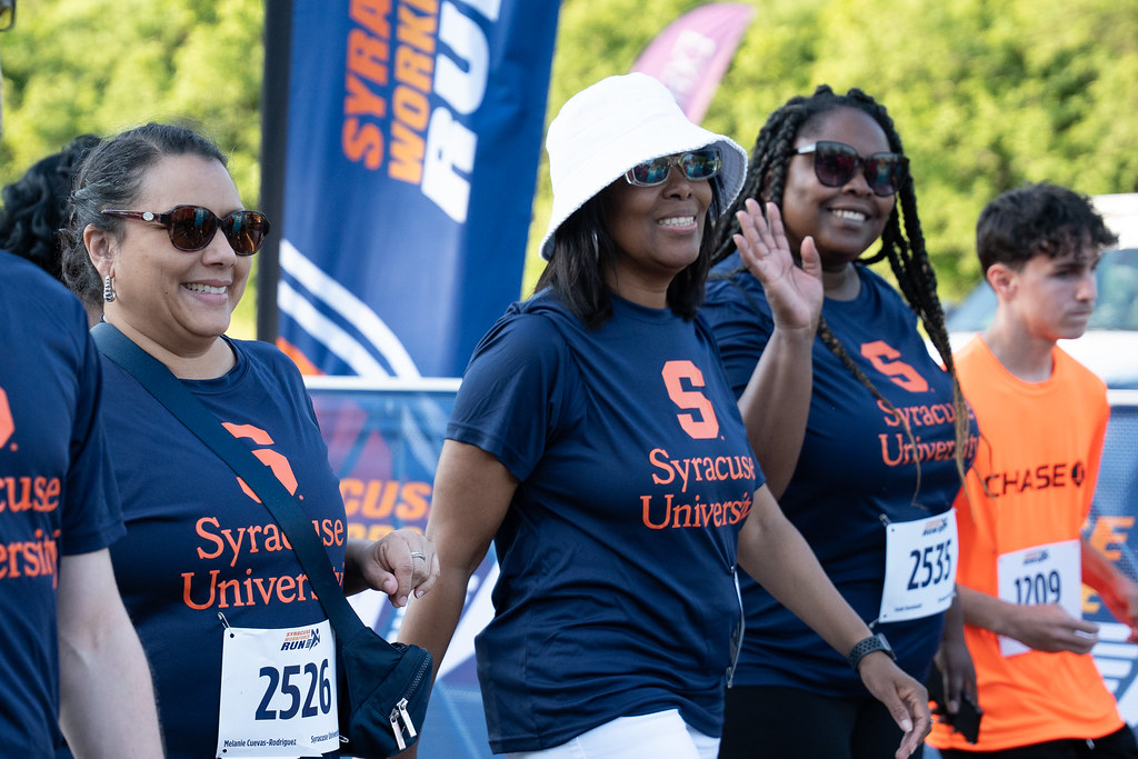 A group of people participate in the Syracuse WorkForce Run, wearing navy blue shirts with 'Syracuse University' and an orange 'S' logo. Visible race bib numbers include 2526, 2533 and 1209. Banners in the background read 'SYRACUSE WORKFORCE RUN.'