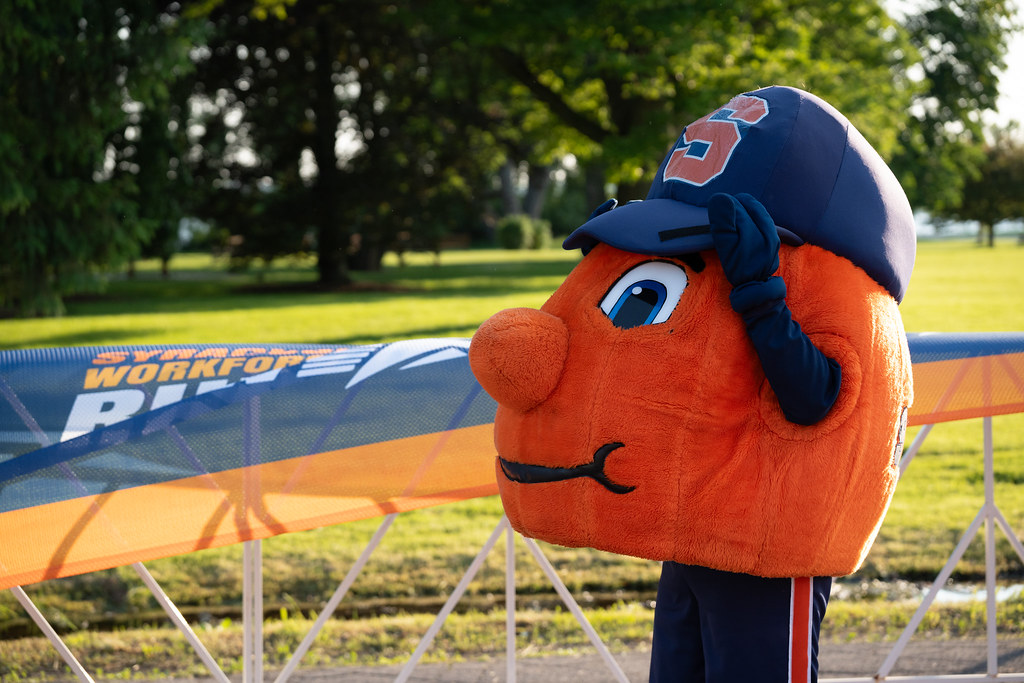 Otto the Orange stands outdoors near a barrier with partially visible text reading 'SYRACUSE WORKFORCE RUN'. Trees and grass are in the background.