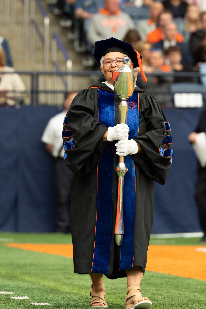 A person in academic regalia holding a ceremonial mace, standing on the field of the JMA Wireless Dome at Commencement 2025. The background includes spectators seated in the stands.