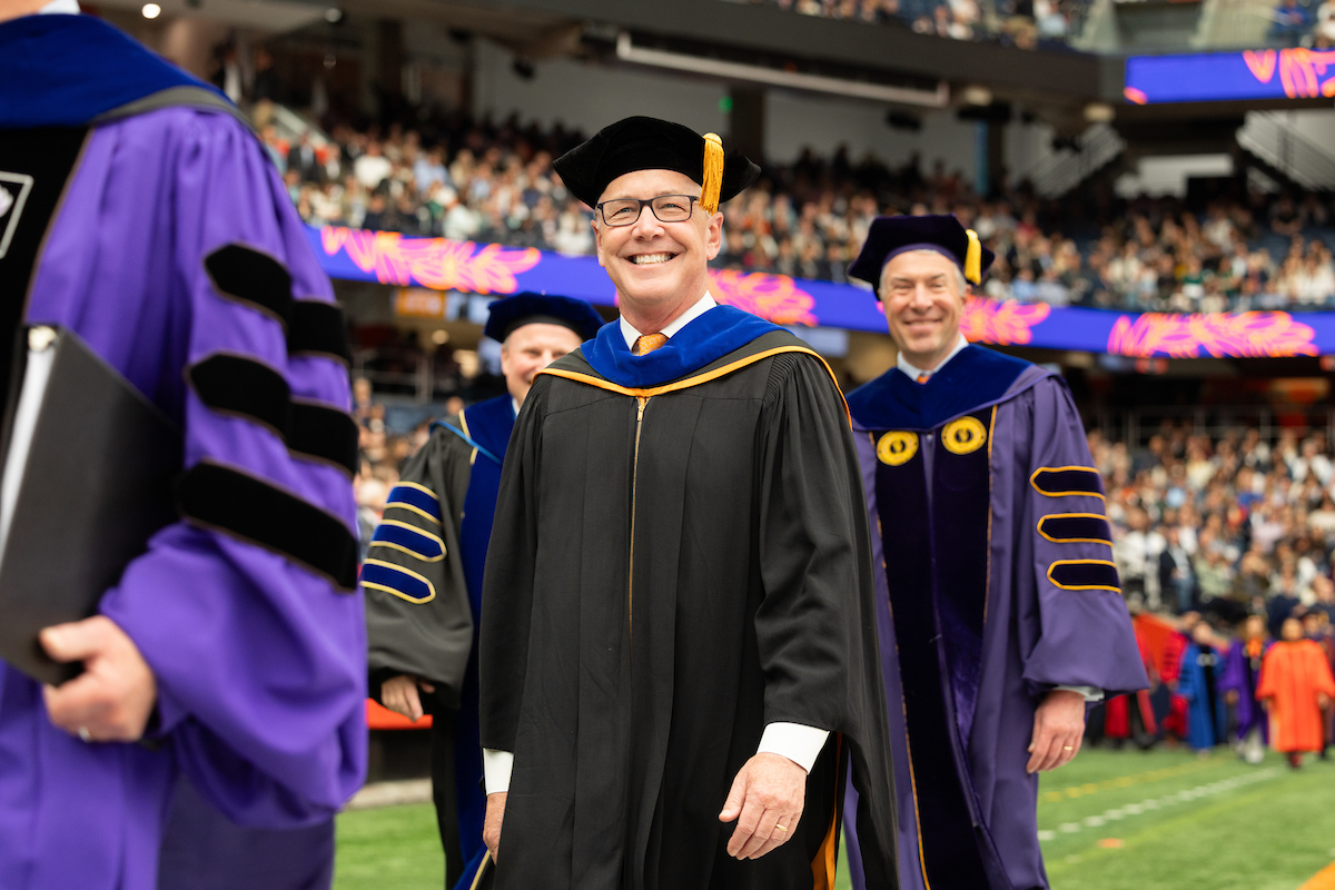 A group of people in academic regalia walk in a procession at Commencement 2025. The individuals are wearing caps and gowns, with some wearing hoods that indicate advanced degrees. The background shows an audience seated in a stadium setting.