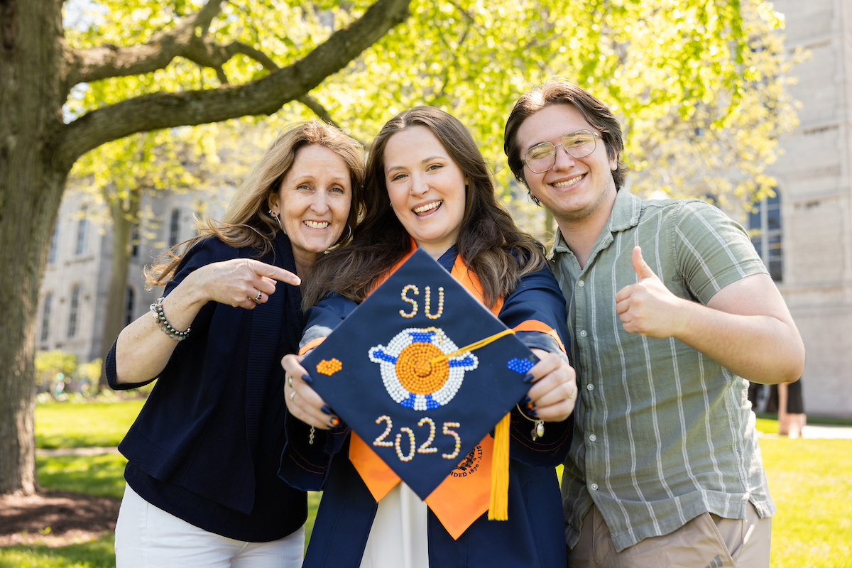 Three people stand outdoors, with trees and a building in the background. The person in the center is holding a decorated graduation cap that reads 'SU 2025' with an orange and blue design. The person on the left is pointing at the cap, while the person on the right is giving a thumbs-up gesture.