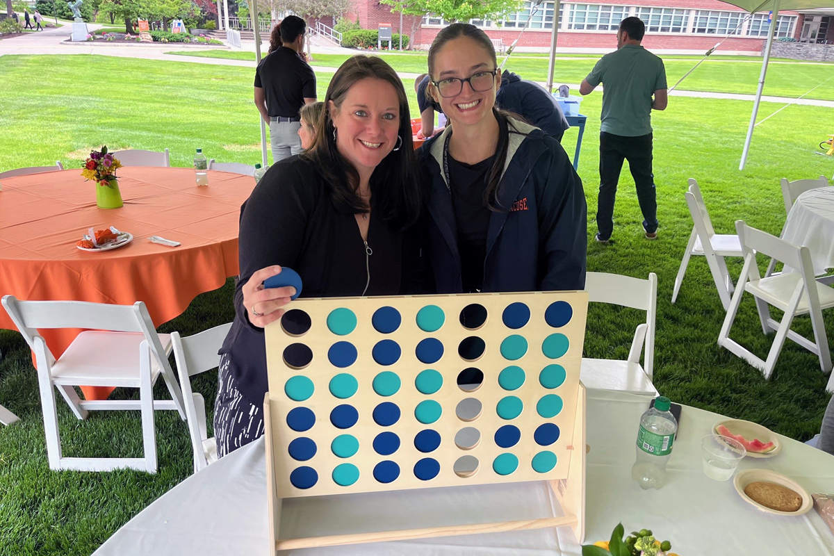 Two people standing behind a large Connect Four game board on a table, with alternating blue and teal discs. The scene is set at an outdoor event featuring white chairs and tables draped with orange and white tablecloths. Vases with flowers, a water bottle, and food items are visible on the tables. In the background, more people are gathered under a tent on a grassy area.