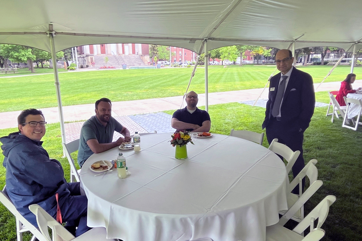 Four people seated around a round table under a white tent on a grassy area, with one person in formal attire standing nearby. The table is covered with a white tablecloth and features a small flower arrangement in the center, along with plates of food and drinks. In the background, an open grassy field with trees and buildings is visible.