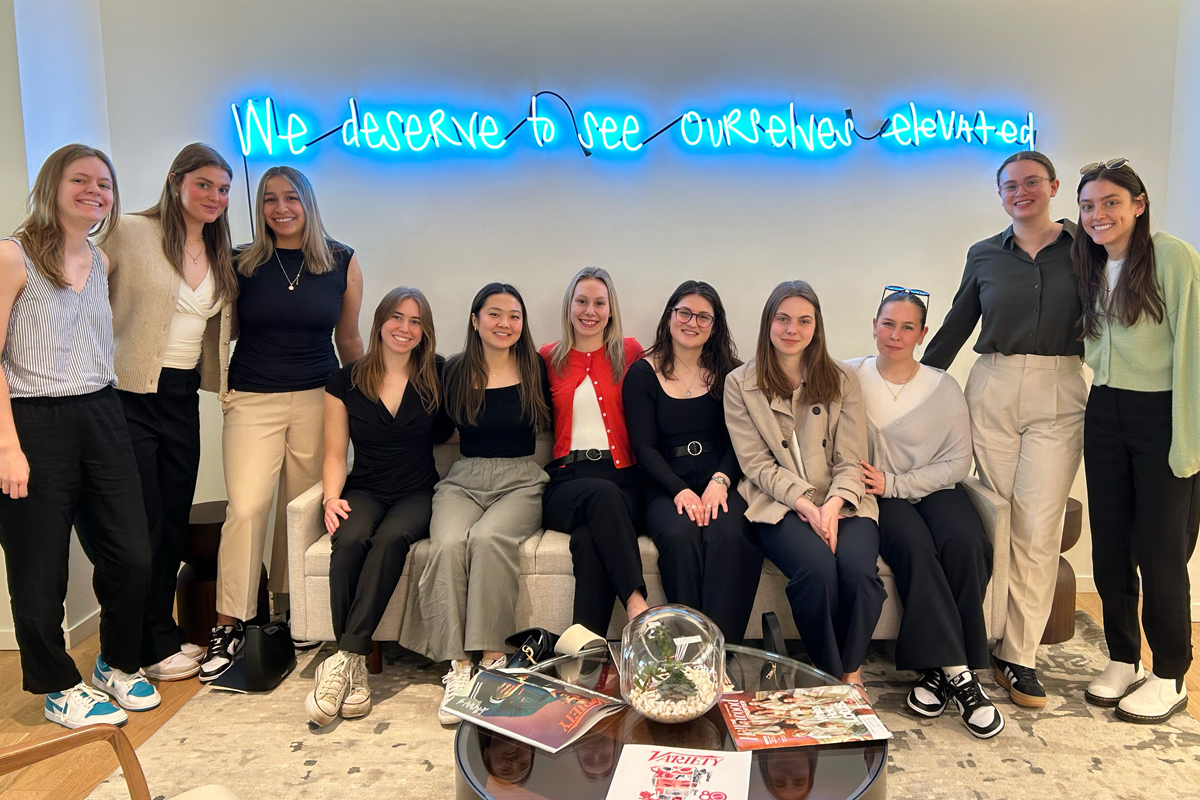 A group of twelve people posing in front of a wall with a neon sign that reads, 'We deserve to see ourselves elevated.' Some individuals are seated on a couch, while others stand behind it. They are dressed in casual to business-casual attire. A coffee table in front of the couch holds magazines and decorative items.