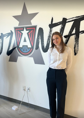 A person standing in front of a wall featuring a bold, stylized graphic design with the word 'ATLANTA' and a large 'A' inside a red and blue shield-like emblem. An electrical outlet with two cords plugged in is visible near the bottom left corner.